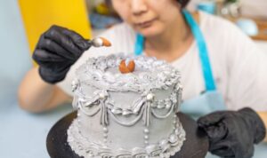 A person wearing black gloves decorates a customised cake Singapore - My Bake Studio style, adding intricate icing to the gray frosted cake and finishing with a small almond-shaped decoration on top.